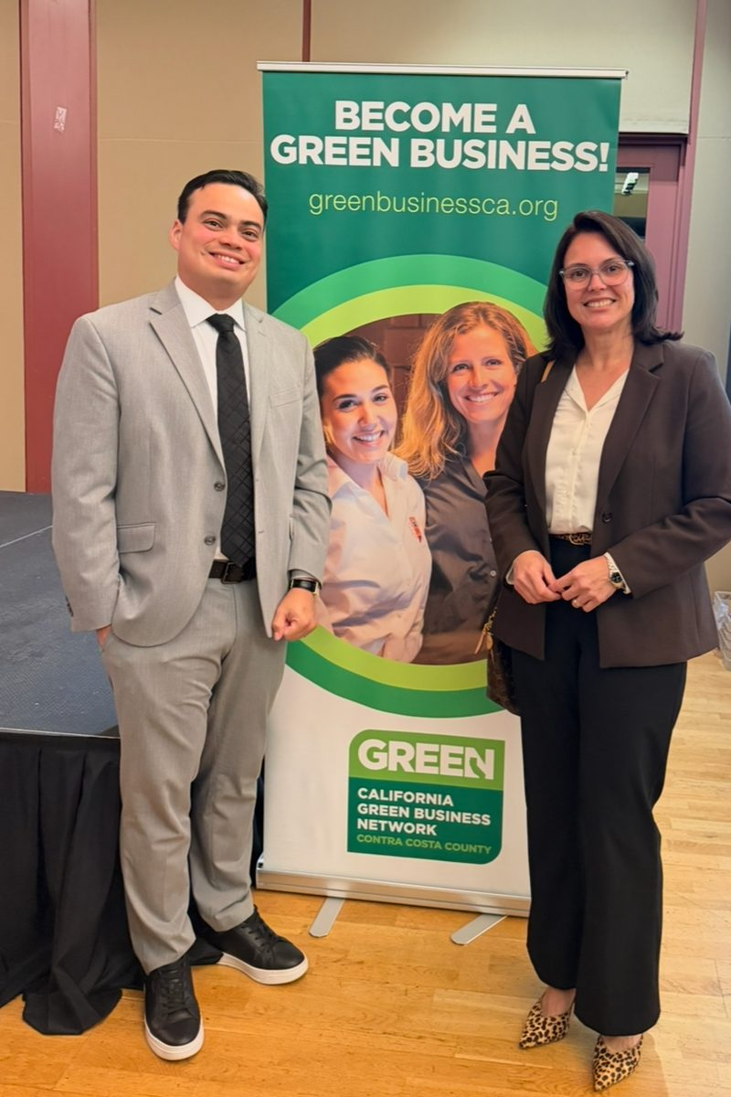 Royal Glow co-founders Diego Coutinho, in a grey suit, and Alessandra Coutinho, in a dark blazer, smiling beside a California Green Business Network, Contra Costa County banner that reads "Become a Green Business!"